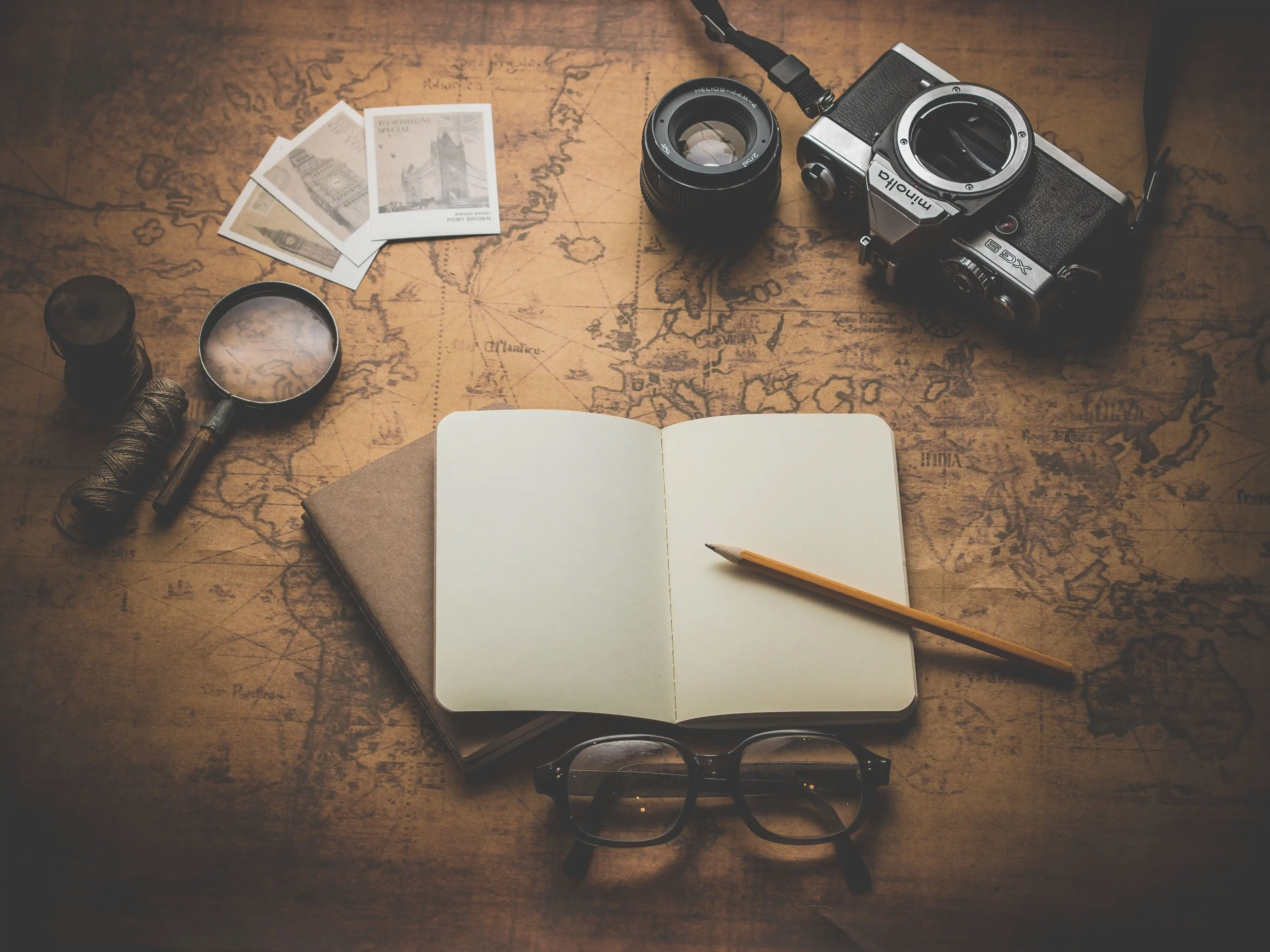 Overhead view of a desk with an old map laid out, an open journal with blank pages resting on top, a vintage camera, a magnifying glass, postcards and a pair of spectacles.